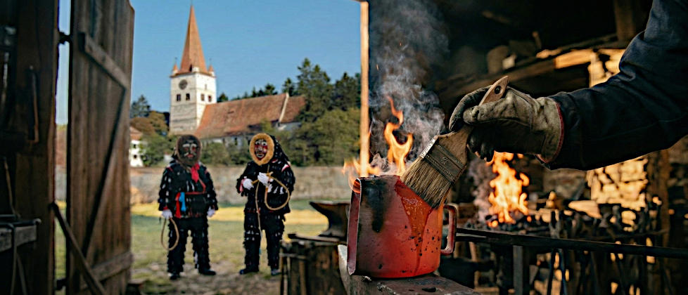 Eine rotglühende Urzel-Schelle wird in einer Schmiede mit einem Pinsel mit Leinöl bestrichen. Im Hintergrund stehen traditionell verkleidete Urzeln vor der historischen Wehrkirche von Cincu.