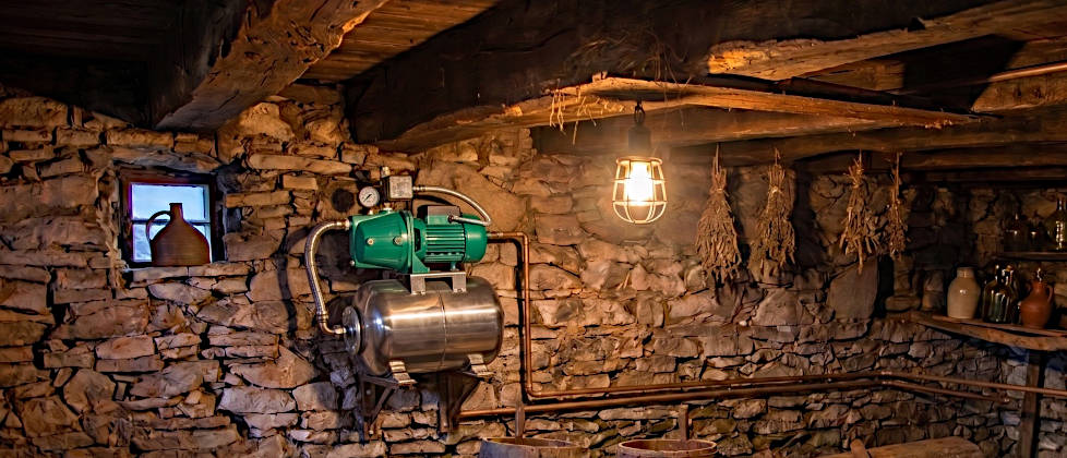Ein modernes Hauswasserwerk mit grüner Pumpe und Edelstahl-Druckkessel, installiert an einer alten Bruchsteinwand in einem rustikalen Keller.