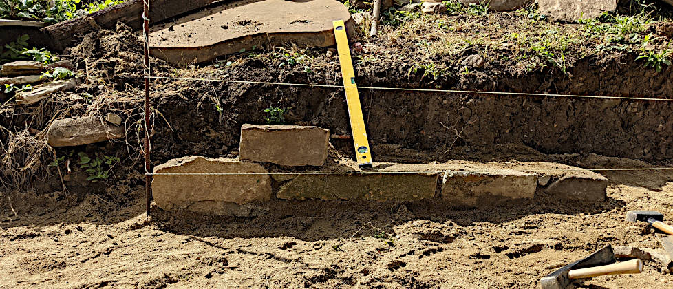 Aufbau einer Trockenmauer aus Feldsteinen an einem Hang: Die erste Steinreihe wird auf einem Sandbett mit einer gelben Wasserwaage und einer Richtschnur exakt ausgerichtet. Im Vordergrund liegen zwei Steinhammer im Sand.