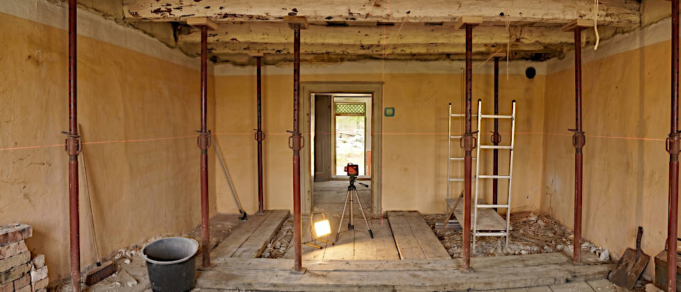 Temporäres Abfangen alter Deckenbalken aus Eiche im Altbau mit lotrecht aufgestellten Stahl-Baustützen auf lastverteilenden Bohlen.
