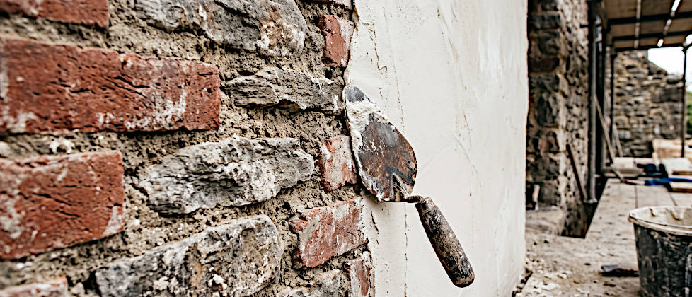Eine teils verputzte historische Wand aus Bruchstein und Feldbrandziegeln, die zeigt, wie man fachgerecht Kalkputz auf Mischmauerwerk auftragen kann, mit einer Maurerkelle im Vordergrund.