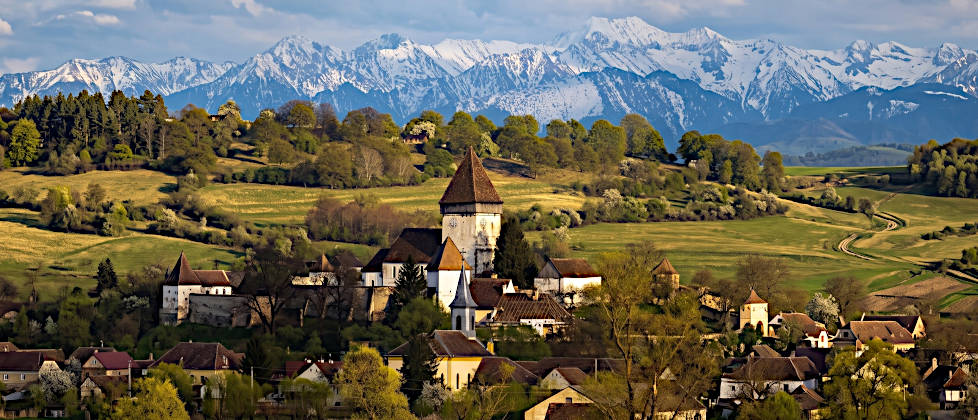 Panoramablick auf die befestigte Kirchenburg Hosman in Siebenbürgen, Rumänien, umgeben von einem malerischen Dorf und sanften grünen Hügeln, mit den vollständig schneebedeckten Gipfeln der Făgăraș-Berge im Hintergrund unter einem teilweise bewölkten blauen Himmel.
