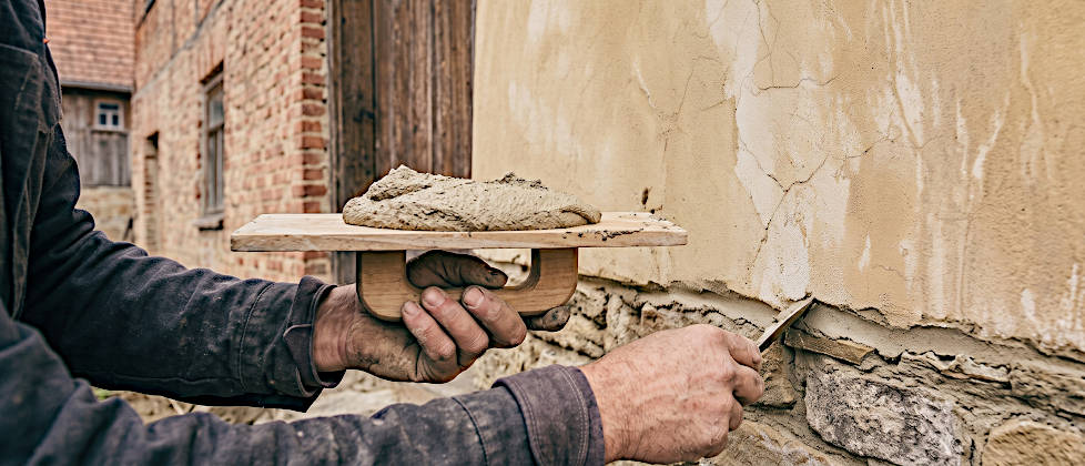 Nahaufnahme der Hände eines Maurers, der die Fugen eines Bruchsteinsockels saniert. In der linken Hand hält er ein waagerechtes Reibebrett mit frischem Kalkmörtel. Mit der rechten Hand drückt er den Mörtel mit einem Fugeisen in die Steinfuge unterhalb einer hell ockerfarben verputzten Hauswand.